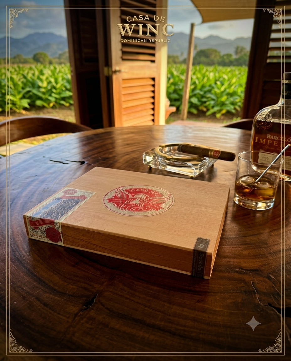 Casa de Wing cigar box on rustic wood table with lit cigar, rum, and Dominican Republic tobacco field in background
